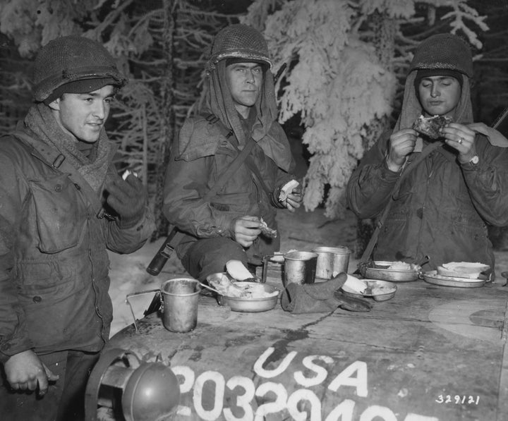 Three soldiers in winter gear eat atop a military vehicle in the snow, with metal cups and plates—a World War II scene reminiscent of Band of Brothers. From "Dying Hard" by Schiffer Publishing, Ltd.