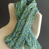 A chunky, hand-knitted scarf made with Schiffer Publishing, Ltd.'s Japanese Paper Yarn in blue and green is draped over a modern, cream chair, set against a plain dark background.