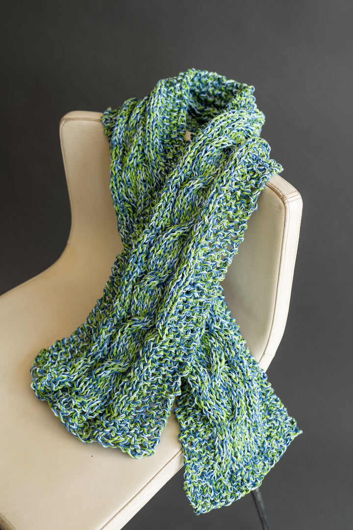 A chunky, hand-knitted scarf made with Schiffer Publishing, Ltd.'s Japanese Paper Yarn in blue and green is draped over a modern, cream chair, set against a plain dark background.