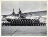 A large group of uniformed personnel poses before a twin-engine seaplane, reflecting US Navy history—an image featured in King's Navy by Schiffer Publishing, Ltd. The group gathers on the tarmac beneath the plane's wings and propellers.