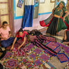Four women in traditional attire, representing Indian artisans, craft and converse among vibrant embroidered textiles. Artisans by Design by Schiffer Publishing, Ltd. captures their creativity in a room full of colorful handmade beauty.