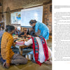 A group of adults and children gather around a low table at Somaiya Kala Vidya as a woman in blue assists, celebrating traditional artisans of India, featured in "Artisans by Design" from Schiffer Publishing, Ltd. A whiteboard is seen behind them.