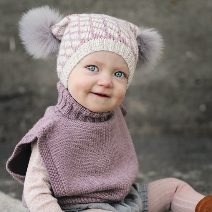 A happy baby with blue eyes wears knits from "Winter Knitting for Little Sweethearts" by Schiffer Publishing, Ltd., including a pink and cream hat with pom-poms, a purple sweater, and a gray skirt, against a blurred gray background.