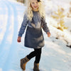 A young child wearing a Nordic-style grey dress from "Winter Knitting for Little Sweethearts" by Schiffer Publishing, Ltd. walks on a snowy path, dressed with black leggings, brown boots, and a black hair bow amid winter trees.