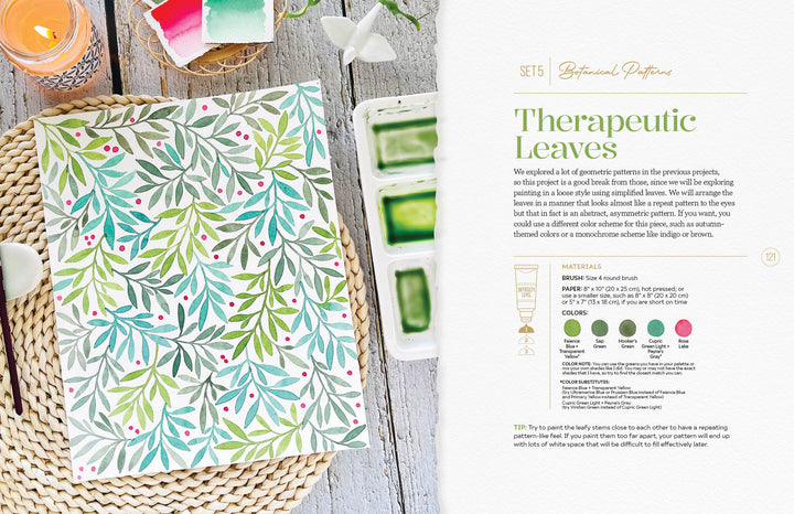 The Modern Watercolor Workshop by Schiffer Publishing, Ltd. lies open on a woven placemat, displaying botanical watercolor leaves with red berries—ideal for beginners. Nearby are paint, a green brush, candle, and towel; “Therapeutic Leaves” is shown on the right page.