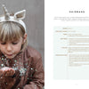 A young child with light hair, wearing a brown jacket and the Magical Crochet unicorn headband from Schiffer Publishing, Ltd., blows glitter into the air against a soft, neutral background.