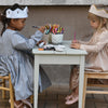 Two young girls wearing crowns and dresses draw with colored pencils at a small table, surrounded by art supplies and Magical Crochet accessories from Schiffer Publishing, Ltd., as they focus on their creative activity.