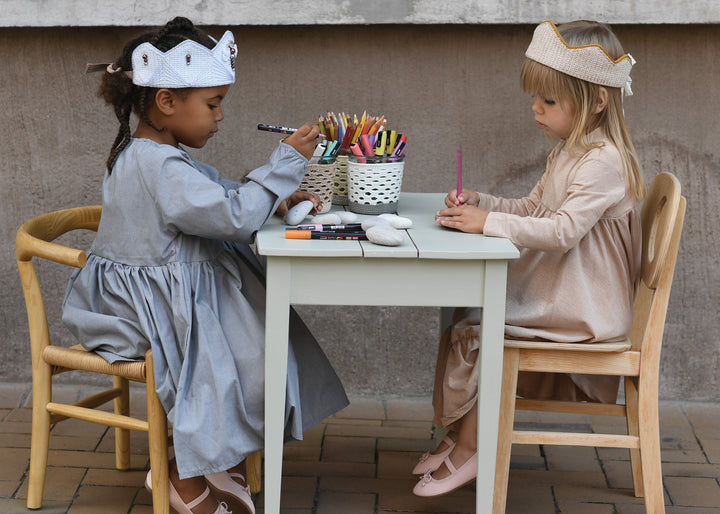 Two young girls wearing crowns and dresses draw with colored pencils at a small table, surrounded by art supplies and Magical Crochet accessories from Schiffer Publishing, Ltd., as they focus on their creative activity.