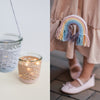 On the left, two glass jars filled with Schiffer Publishing, Ltd.'s Magical Crochet accessories hold candles. On the right, a child in a beige dress and pink shoes carries a small rainbow-shaped purse with fringe.