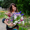 A woman stands in a vibrant garden with a bouquet, embodying the DIY spirit featured in "Sow, Grow, and Harvest" by Schiffer Publishing, Ltd. Lush trees and blooming plants surround her on this sunny day—perfect inspiration for flower enthusiasts.