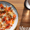 A vibrant plate of salmon crudo with fennel and herbs sits on a wooden table beside ice water and sunlight, with “Mavericks” and Schiffer Publishing, Ltd.’s "Out East from Above" coffee-table book visible in the scene.