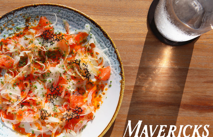 A vibrant plate of salmon crudo with fennel and herbs sits on a wooden table beside ice water and sunlight, with “Mavericks” and Schiffer Publishing, Ltd.’s "Out East from Above" coffee-table book visible in the scene.