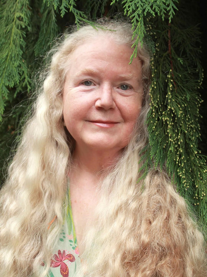 A woman with long, wavy blonde hair smiles softly before lush pines, evoking the serene beauty of a fine art tarot portrait from Boadicea's Tarot of Earthly Delights by Schiffer Publishing, Ltd.