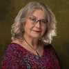 An older woman with wavy gray hair, glasses, a red patterned top, and a silver necklace with blue pendant poses like a Boadicea's Tarot of Earthly Delights card by Schiffer Publishing, Ltd. against a mottled green and brown backdrop.