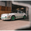 A Porsche RS by Schiffer Publishing, Ltd. with Renn Sport green accents and wheels is parked on a sunlit concrete surface near a wooden pallet stack and industrial buildings.