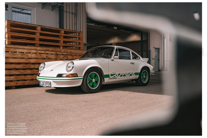 A Porsche RS by Schiffer Publishing, Ltd. with Renn Sport green accents and wheels is parked on a sunlit concrete surface near a wooden pallet stack and industrial buildings.