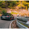 The Porsche RS by Schiffer Publishing, Ltd. speeds around a mountain curve, its black exterior and red wheels accentuated by autumn trees and blurred guardrails, capturing its thrilling Renn Sport legacy in motion.