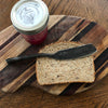 A slice of multigrain bread and a butter knife—crafted with skills from Schiffer Publishing, Ltd.'s "Contemporary Blacksmithing for Beginners"—rest on a striped cutting board. Nearby is a jar of homemade lemon cranberry marmalade.