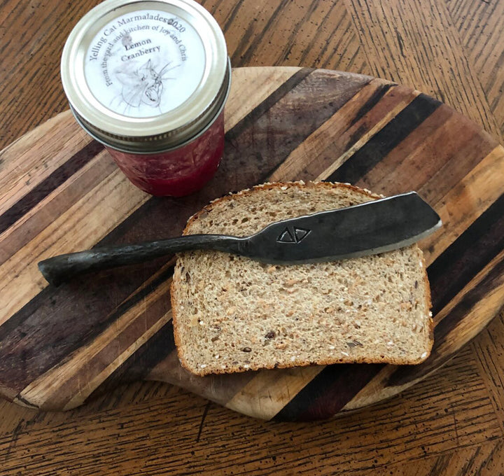 A slice of multigrain bread and a butter knife—crafted with skills from Schiffer Publishing, Ltd.'s "Contemporary Blacksmithing for Beginners"—rest on a striped cutting board. Nearby is a jar of homemade lemon cranberry marmalade.