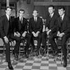 Five men in dark suits and ties sit on bar stools at a counter in a retro-style setting with a checkered floor, evoking the timeless atmosphere featured in Schiffer Publishing’s Richard Manuel.