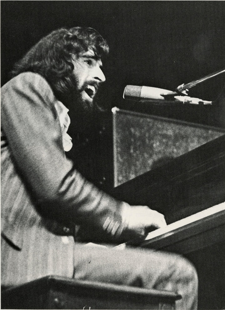 A bearded man with long hair passionately sings into a microphone while playing piano, evoking the legendary rock music captured in "Richard Manuel" by Schiffer Publishing, Ltd., as seen in this expressive black and white photo.