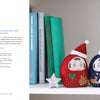 The book "Kawaii Appliqué Quilts from Japan" by Schiffer Publishing, Ltd. rests on a white shelf with books, plants, and silver stars, creating a festive and cozy holiday display inspired by Japanese quilting.