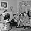 A group of five in 1940s attire converse in a living room with papers, reminiscent of scenes from "The Art of Classic Crime and Mystery Movies" by Schiffer Publishing, Ltd., featuring elegant drapes, a painting, and classic intrigue.