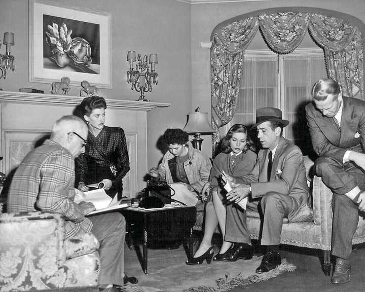 A group of five in 1940s attire converse in a living room with papers, reminiscent of scenes from "The Art of Classic Crime and Mystery Movies" by Schiffer Publishing, Ltd., featuring elegant drapes, a painting, and classic intrigue.