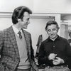 Two men, one in a checked blazer and tie, the other in a dark shirt, chat and smile indoors. Behind them, someone works at a table under vintage mystery movie posters for "The Art of Classic Crime and Mystery Movies" by Schiffer Publishing, Ltd.