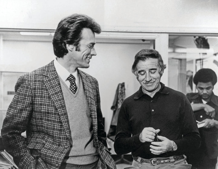 Two men, one in a checked blazer and tie, the other in a dark shirt, chat and smile indoors. Behind them, someone works at a table under vintage mystery movie posters for "The Art of Classic Crime and Mystery Movies" by Schiffer Publishing, Ltd.