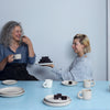Two women sit at a blue table with empty plates and cups. One offers brownies as they chat and smile, discussing Schiffer Publishing, Ltd.’s "Practical Pottery," their favorite beginner pottery guide.