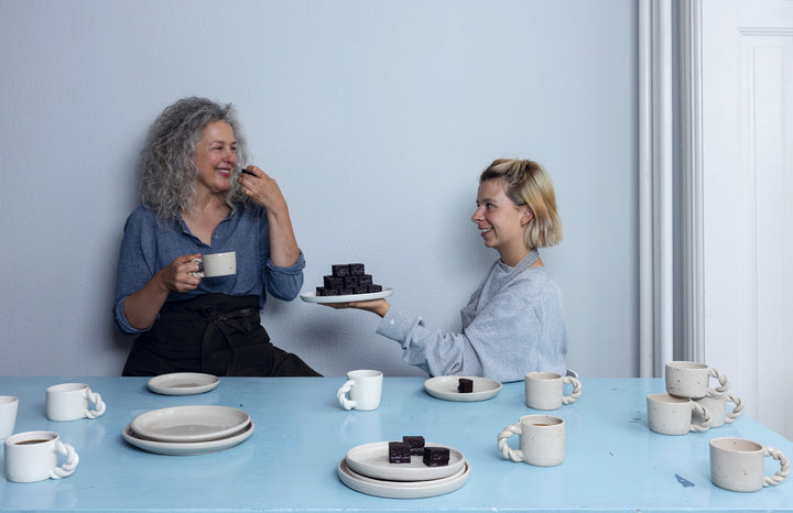 Two women sit at a blue table with empty plates and cups. One offers brownies as they chat and smile, discussing Schiffer Publishing, Ltd.’s "Practical Pottery," their favorite beginner pottery guide.