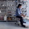 In a bright studio filled with ceramic bowls, a woman uses a pottery wheel. The text "Practical Pottery" from Schiffer Publishing, Ltd. appears, ideal for beginners seeking an essential pottery guide.