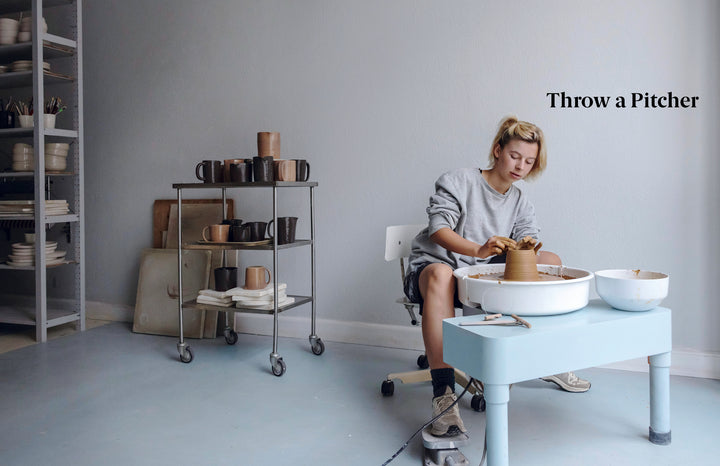 A woman creates pottery on a wheel, shaping clay into a pitcher, with finished ceramics displayed nearby. "Throw a Pitcher" is on the wall. Practical Pottery by Schiffer Publishing, Ltd. inspires the creative scene.