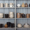 Schiffer Publishing, Ltd.’s “Practical Pottery” displays ceramic mugs, bowls, cups, and vases in neutral tones—wheel-thrown and arranged on grey wooden shelves against a light paneled wall for a balanced look.