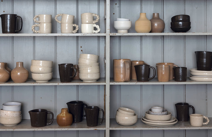 Schiffer Publishing, Ltd.’s “Practical Pottery” displays ceramic mugs, bowls, cups, and vases in neutral tones—wheel-thrown and arranged on grey wooden shelves against a light paneled wall for a balanced look.