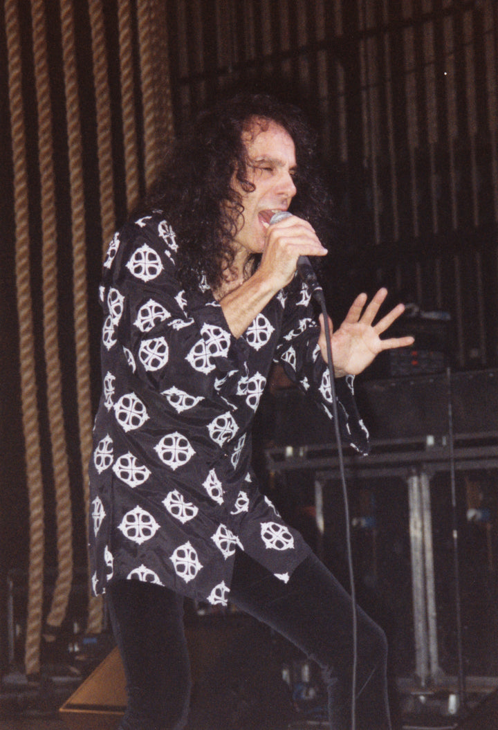 A singer with long curly hair, reminiscent of Ronnie James Dio, passionately performs on stage wearing a black shirt with a white skull, as seen in Schiffer Publishing, Ltd.’s "DIO: The Unholy Scriptures." Stage equipment and ropes are visible in the background.