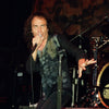 A male rock singer with long curly hair channels Ronnie James Dio while performing on stage, promoting "DIO: The Unholy Scriptures" from Schiffer Publishing, Ltd., with drums and speakers in the background.
