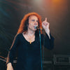 A man with long curly hair, in a black shirt and cross necklace, performs on stage under vibrant lights—capturing the spirit of Ronnie James Dio, as featured in "DIO: The Unholy Scriptures" from Schiffer Publishing, Ltd.