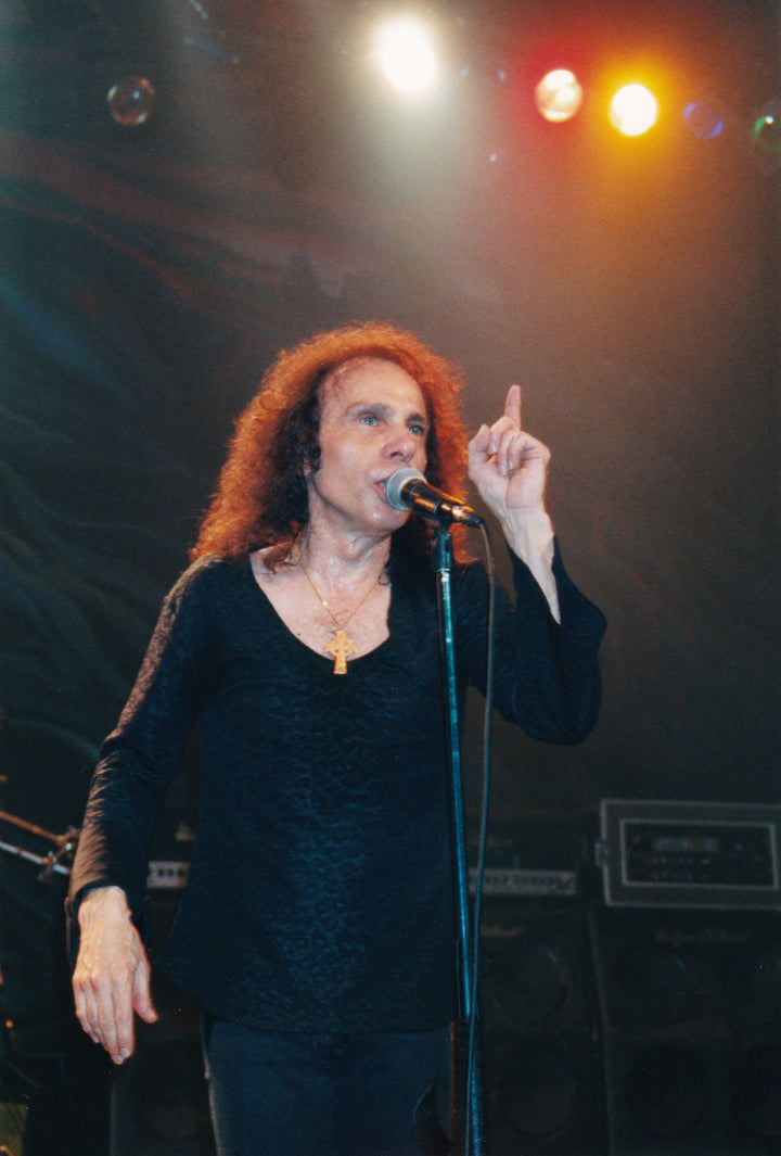 A man with long curly hair, in a black shirt and cross necklace, performs on stage under vibrant lights—capturing the spirit of Ronnie James Dio, as featured in "DIO: The Unholy Scriptures" from Schiffer Publishing, Ltd.