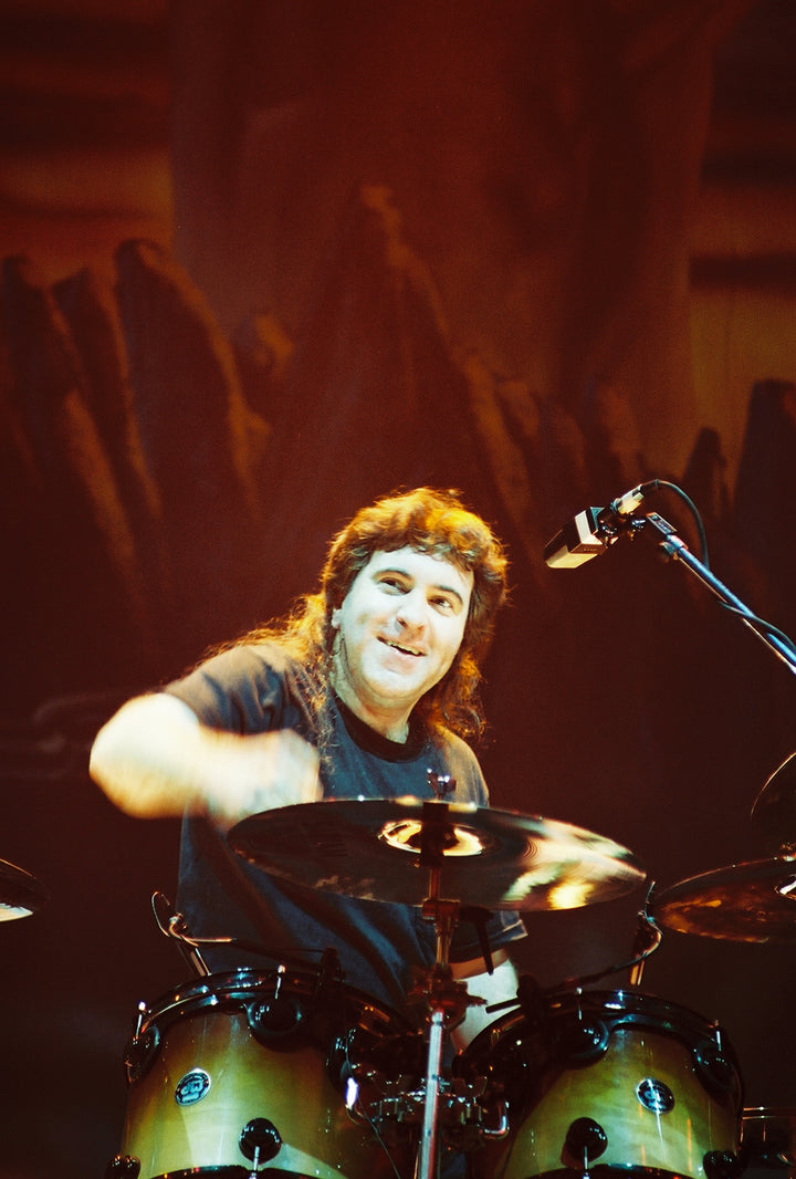 A drummer with long dark hair plays onstage, smiling as he raises a drumstick, capturing the energetic vibe of Dio—perfect for fans of DIO: The Unholy Scriptures from Schiffer Publishing, Ltd.