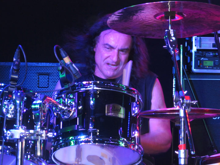 A drummer with long dark hair rocks out on stage amid colorful lights and gear, evoking DIO's iconic Holy Diver era—captured in DIO: The Unholy Scriptures by Schiffer Publishing, Ltd.