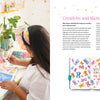 An artist at a desk paints vibrant flowers in the Creative Sketchbook Magic by Schiffer Publishing, Ltd. The right page features a watercolor with the words: Your wings already exist. All you have to do is fly—inspiring daily creativity.