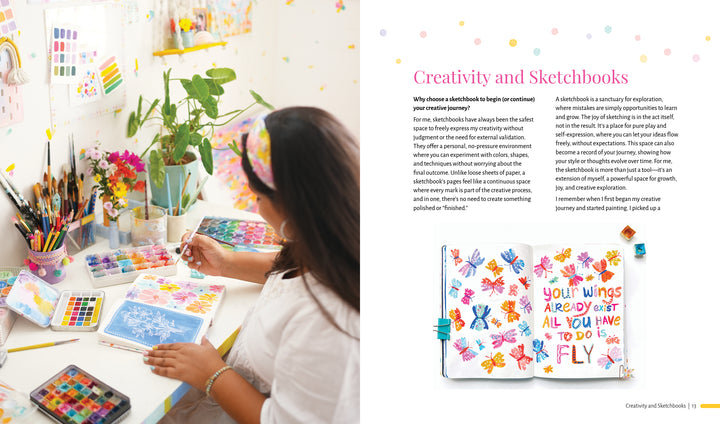 An artist at a desk paints vibrant flowers in the Creative Sketchbook Magic by Schiffer Publishing, Ltd. The right page features a watercolor with the words: Your wings already exist. All you have to do is fly—inspiring daily creativity.
