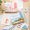 Schiffer Publishing, Ltd.'s Watercolor the Easy Way Paint-Your-Own Watercolor Cards Flowers kit is arranged on a light wooden surface with brushes, paper featuring floral designs, a striped cloth, plant, flower, notecards, and a blue envelope. "Collect Both Kits!" label shown.