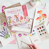 The Schiffer Publishing, Ltd. Watercolor the Easy Way Paint-Your-Own Watercolor Cards Flowers kit is shown with painted floral cards, a palette, pink envelopes, and a vase of white flowers beside a hand-painted card.