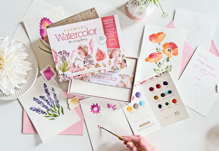 The Schiffer Publishing, Ltd. Watercolor the Easy Way Paint-Your-Own Watercolor Cards Flowers kit is shown with painted floral cards, a palette, pink envelopes, and a vase of white flowers beside a hand-painted card.