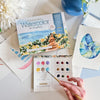 A hand holds a paintbrush over Schiffer Publishing, Ltd.'s Watercolor the Easy Way Paint-Your-Own Watercolor Cards Coastal kit, surrounded by ocean-themed watercolor paintings, a large white flower, and a blue vase.