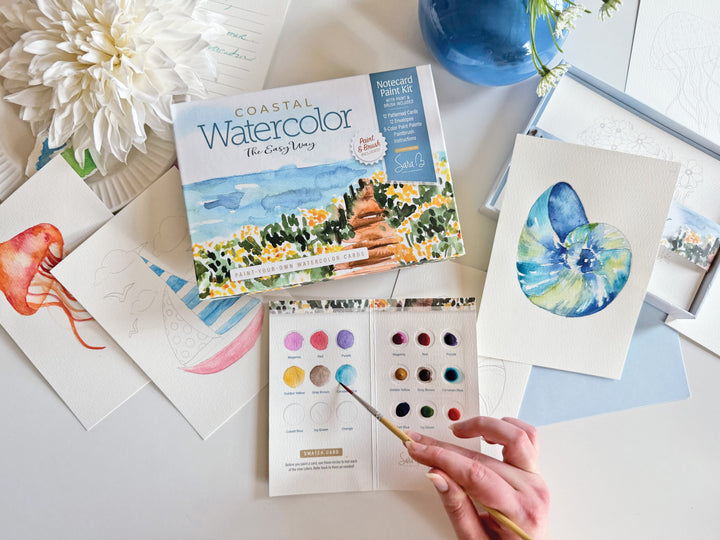 A hand holds a paintbrush over Schiffer Publishing, Ltd.'s Watercolor the Easy Way Paint-Your-Own Watercolor Cards Coastal kit, surrounded by ocean-themed watercolor paintings, a large white flower, and a blue vase.