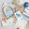 A table displays Schiffer Publishing’s "Watercolor the Easy Way Paint-Your-Own Watercolor Cards Coastal" kit, with paint samples, brushes, blank and painted notecards (jellyfish, seashell), a palette, and a white flower in a blue vase.
